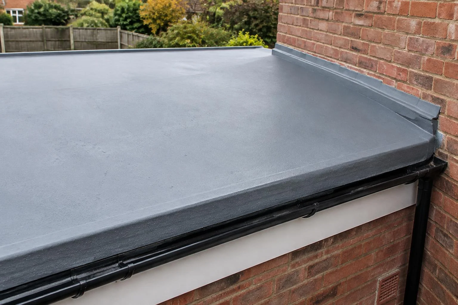 Grey seamless flat-roof edge, gutter and brick wall in a UK residential setting. AI-generated illustration, not an LMB project.